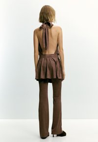 Ženska stoji s hrbtom videnim, oblečena v rjavo halterneck bluzo zvezano na vratu, ujemajoče se široke hlače in kratko krilo, plastčeno preko hlač.
