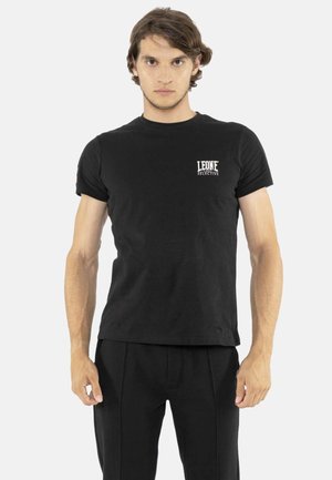 Man staat recht vooruit kijkend, draagt een zwart T-shirt met korte mouwen met het "Leone Selective"-logo op de borst en een zwarte broek.
