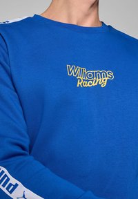 Niebieska koszula z długim rękawem z żółtym logo "Williams Racing" na piersi i białym brandingiem Puma na rękawach. Gładka tekstura.