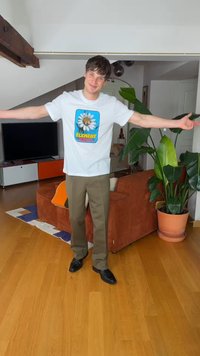 Mladý muž v bielom tričku s grafickým motívom a hnedých nohaviciach stojí s rukami rozpaženými v obývacej izbe s rastlinami a nábytkom.