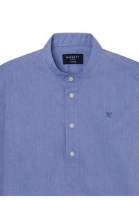 Camisa de chambray azul claro con cuello de banda, cinco botones y un pequeño logo bordado en el lado izquierdo del pecho. Textura suave y diseño casual.