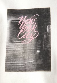 Camiseta gráfica con un estampado en blanco y negro que presenta el texto "New York City" en cursiva rosa, sobre un fondo de tela de color claro.