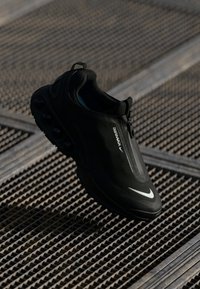 Melnas Nike Air Max kedas ar rāvējslēdzēju un redzamu gaisa spilvenu zolīti uz teksturētas metāla režģa virsmas.