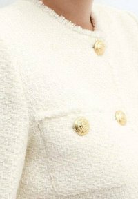 Veste blanche texturée avec deux boutons dorés et une poche avant ornée d'un bouton similaire. Bords effilochés au col.