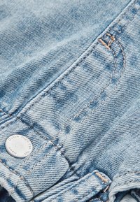 Gros plan d'un tissu denim bleu clair montrant des coutures détaillées et un bouton en métal au bord d'une poche.