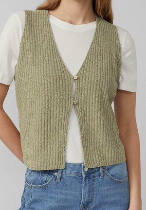 Femme portant un gilet côtelé vert olive clair boutonné sur un T-shirt blanc à manches courtes et un jean bleu, debout avec les mains derrière le dos.