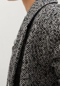 Detailní záběr osoby nosící černobílý kabát s vzorem rybí kostí a širokým límcem.