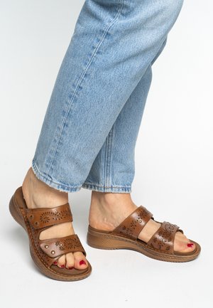 Brown leather sandals with perforated design, dual adjustable straps, and a textured platform sole, paired with light blue jeans.