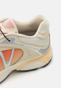 Baskets de sport beiges avec une tige en maille texturée, accents orange et détails gris. Présente une semelle rembourrée et un design léger.
