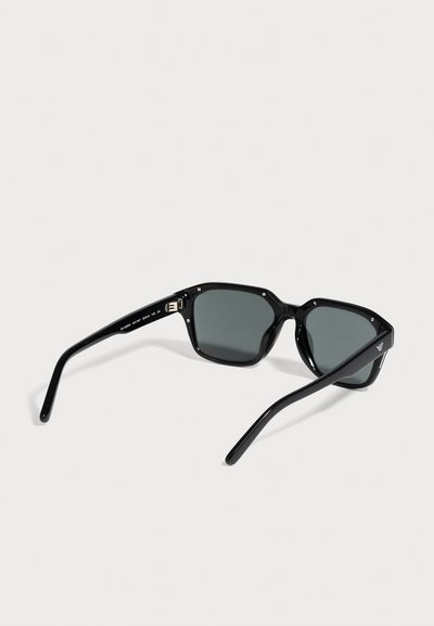 Lunettes de soleil noires rectangulaires aux verres teintés foncés, représentées de biais sur un fond uni clair.