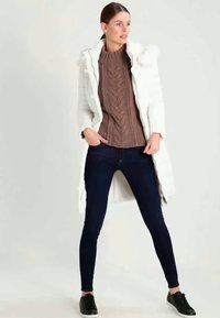 Mulher vestindo um longo casaco acolchoado branco sobre um suéter de malha marrom, jeans skinny escuros e ténis pretos, em pé com as mãos nos bolsos.