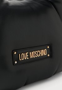 Schwarze Handtasche mit glatter Textur; verfügt über eine vergoldete Logo-Plakette und Nieten-Details als dekorative Akzente.