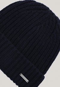 Granatowa dziergana czapka z prążkowanym wzorem, z zawiniętym mankietem i srebrną metalową metką z napisem "PROCHNIK" po boku.