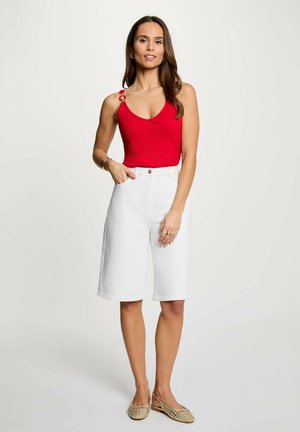 Donna che indossa un top rosso senza maniche con dettaglio anello dorato, pantaloncini bianchi fino al ginocchio e scarpe piatte beige intrecciate, in piedi davanti a uno sfondo semplice.