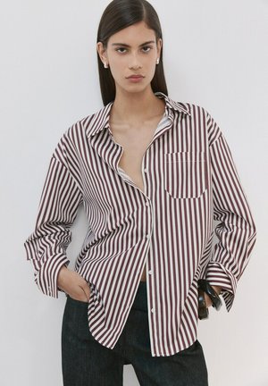Joven con cabello lacio oscuro, vestida con camisa de botones a rayas verticales marrón y blanca y pantalones oscuros, manos en los bolsillos, expresión neutral.