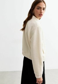 Witte, gestructureerde cropped jack met een staande kraag, zijzakken en geribde manchetten, gedragen met een zwarte kledingstuk. Effen achtergrond.