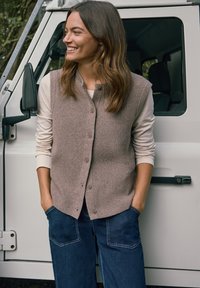 Femme souriante les mains dans les poches en jean, portant un gilet en maille beige boutonné par-dessus une chemise à manches longues, debout près d'une porte de véhicule blanche.