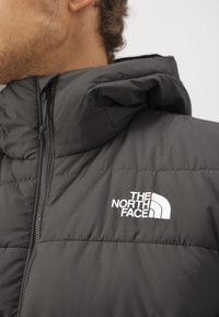 Černá prošívaná bunda se zapínáním na zip a nastavitelnou kapucí. Na hrudi je bílý logo, které zdůrazňuje hladkou, lehkou texturu.