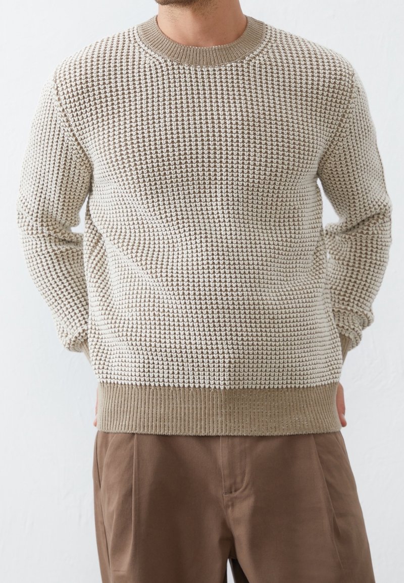 Maglione lavorato a maglia beige chiaro con un motivo testurizzato, dotato di scollo a girocollo e orlo a costine. Indossato con pantaloni marroni.
