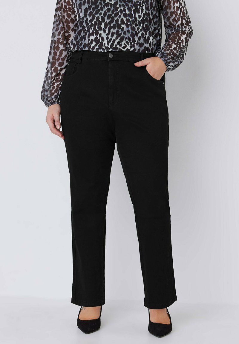 Schwarze Denim-Hosen mit geradem Bein, ausgestattet mit Fronttaschen, hoher Taille und einem Knopfverschluss. Kombiniert mit einer gemusterten Bluse und High Heels.