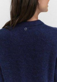 Granatowy, teksturowany sweter z dzianiny z okrągłym dekoltem i małym, srebrnym okrągłym akcentem z tyłu. Miękka, puszysta tkanina.