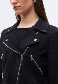 Γυναίκα που φοράει μαύρο υφασμάτινο σακάκι με ασημένια φερμουάρ και κουμπιά πατητά, συνδυασμένο με ασημένια σκουλαρίκια κρίκους και μαύρο τοπ.