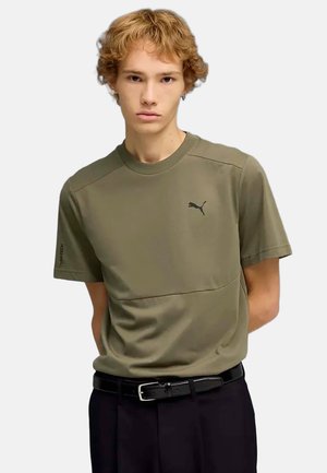 Jonge man met krullend blond haar, gekleed in een olijfgroen Puma T-shirt en zwarte broek met een zwarte riem, staand tegen een effen achtergrond.