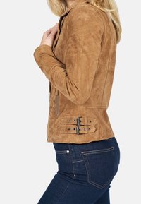 Giacca in suede marrone con un design aderente, zip laterali e dettagli con fibbia in vita. La texture è morbida e leggermente usurata.