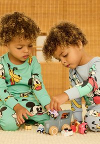 Kinderen dragen kleurrijke pyjama's met tekenfilmfiguren erop, waarbij de één groen en de ander blauw is, omringd door verschillende speelgoedauto's op een zachte ondergrond.