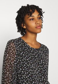 Blusa preta com estampado floral, com mangas longas transparentes, apresentando pequenas flores em tons claros e um decote redondo com um detalhe de franzido sutil.