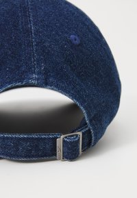 Jeans kapa v temno modri barvi s ukrivljenim robom, nastavljivim trakom in kovinsko zaponko. Ima detajle šivanja ter gumb na vrhu.