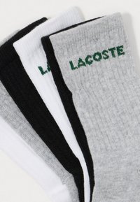 Trzy pary skarpetek damskich w kolorze szarym, czarnym i białym, każda z wyszywanym napisem "LACOSTE" w zielonym kolorze blisko ściągacza.