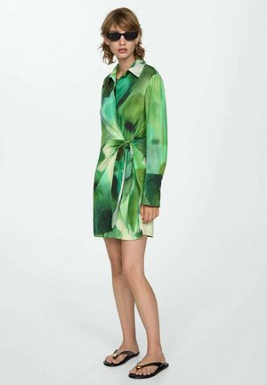 Zelené šaty s kvetinovým vzorom, dlhými rukávmi, viazaným pásom a golierom. Model má na sebe čierne slnečné okuliare a šľapky.