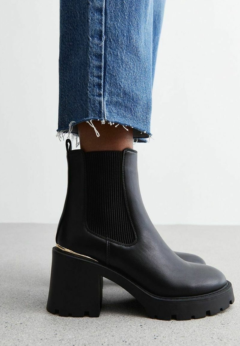 Bottines en cuir noir avec côtés élastiques côtelés, talon bloc épais noir, et garniture accentuée en or. Portées avec un jean court en denim effiloché.
