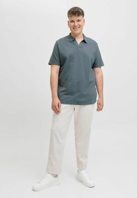 Polo shirt gris foncé avec un col court, tissu texturé, assorti à un pantalon crème et des baskets blanches, se tenant sur un fond uni.