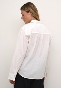 Camisa blanca de botones con mangas largas, que presenta un sutil patrón de pequeños puntos azules y una espalda plisada para un mejor ajuste y movimiento.