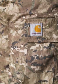Kamouflage-mönstrad tyg med nyanser av brunt och grönt; inkluderar en Carhartt-logopatch i orange på vit bakgrund.
