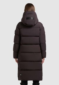 Lange, donkerbruine pufferjack met een capuchon, voorzien van horizontale stiksels en een logo-opdruk nabij de zoom. Gladde textuur, knielange uitvoering.