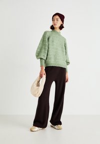 Pull en maille vert clair avec des manches bouffantes, associé à un pantalon large côtelé marron foncé. Tenant un sac à main doux de couleur crème.