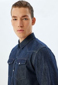 Mörkblå denimskjorta med krage, dubbla bröstfickor med tryckknapp och detaljerad sömnad längs axlarna.