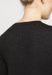 Femme blonde portant un pull en maille gris foncé à col rond, montrant son dos et son profil sur un fond uni.
