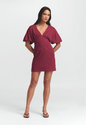 Bordeaux jurk met korte pofmouwen en een diepe V-hals. Heeft een geraffineerd detail bij de buste, een soepele textuur en een lengte boven de knie.