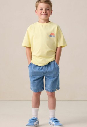 Jongentje met een glimlach en zijn handen in zijn zakken, draagt een geel T-shirt met een zonlogo, blauwe denim shorts, witte sokken en blauwe sneakers.