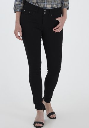 Černé skinny džíny s hladkou texturou, designem pěti kapes a zapínáním na knoflík. Kombinované s černými otevřenými podpatky na světlém pozadí.