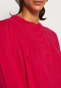 Fuchsia blus, lättviktig tyg, knäppt fram, plisserad design längs axlarna, mandarin-krage, slät struktur och subtil lyster.