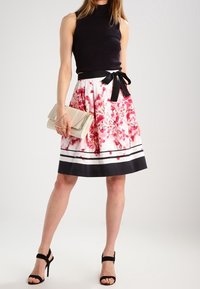 Haut sans manches noir associé à une jupe blanche présentant des motifs floraux roses, une ceinture noire et des rayures blanches au bas. Pochette dorée.