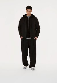 Črna zip-up kapuca z vrvicami, poleg sive majice, kombinirana z ohlapnimi črnimi hlačami in srebrnimi športnimi čevlji. Minimalističen dizajn.