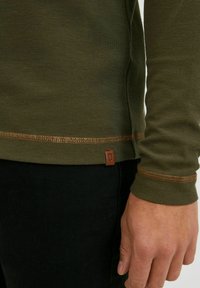 Majica dugih rukava u maslinasto zelenoj boji, izrađena od teksturiranog materijala, sa detaljima od bakrene končane šavove i malom kožnom oznakom na rubu.