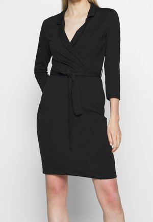 Femme portant une robe portefeuille noire longueur genou avec des manches trois-quarts et une ceinture nouée à la taille, debout devant un fond uni.