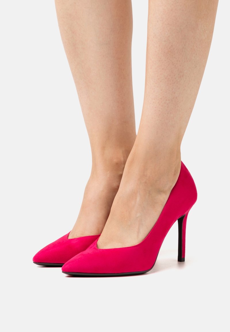 Pieds portant des escarpins en daim rose vif à bout pointu avec des talons aiguilles noirs fins sur un fond blanc.